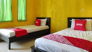 Two single beds with black frames and white bedding, each with a red pillow and a red blanket. The room has yellow walls with some wear and tear, and green curtains on the windows. - OYO 90333 Wisma Tenang Jaya Syariah (Jatiwangi)