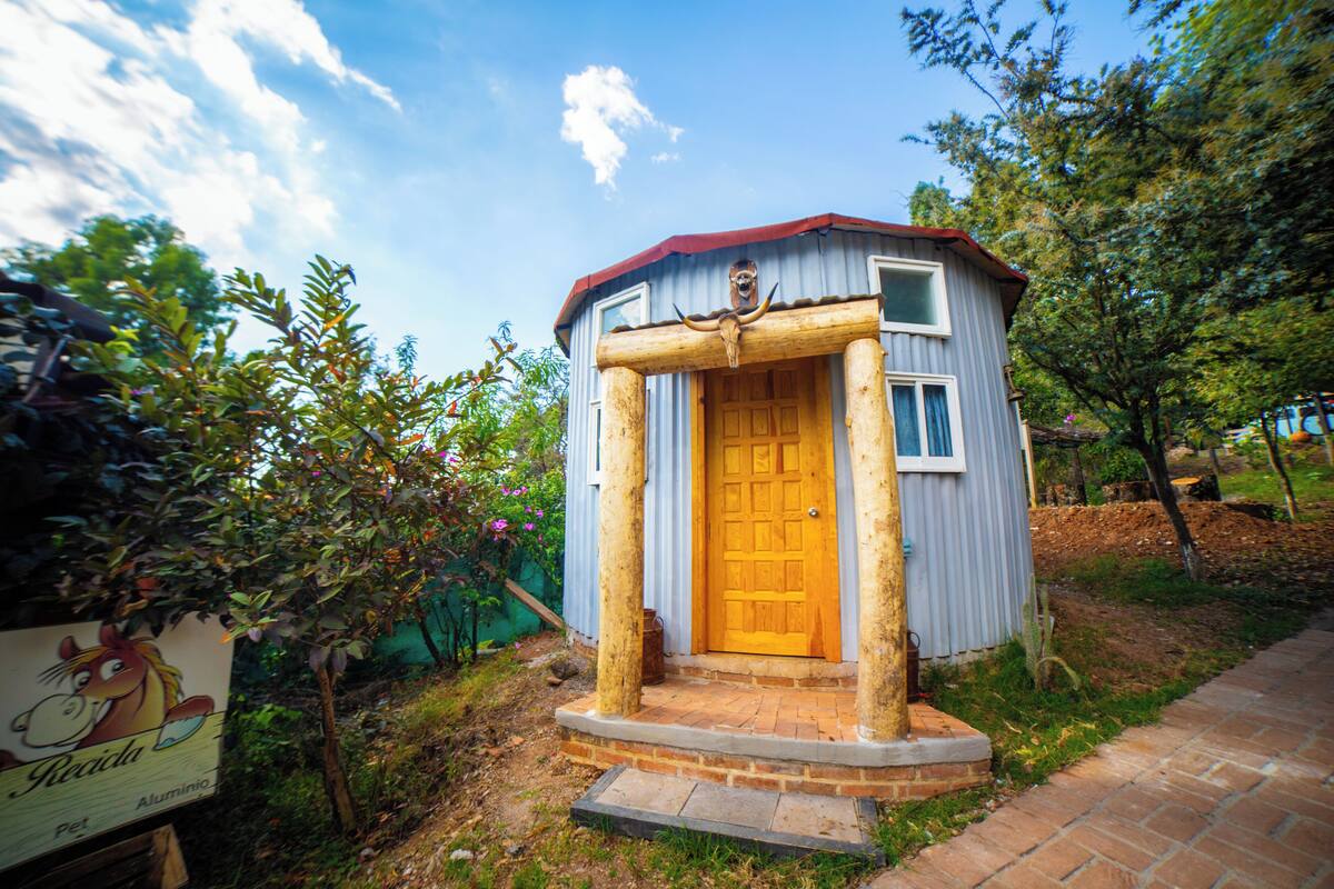 A small wooden cabin with a wooden door and a red roof, surrounded by greenery.