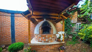 Un horno de ladrillo al aire libre bajo una pérgola de madera, rodeado de macetas con plantas y un muro de ladrillo.