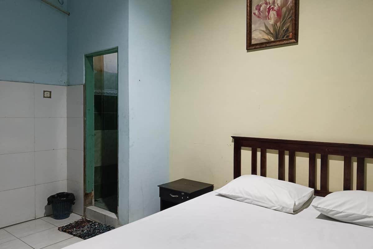 A bedroom with a bed, a nightstand, a trash bin, a bathroom with a shower, and a painting on the wall.