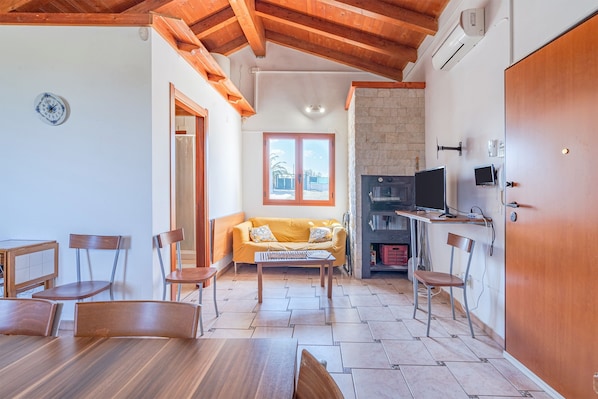 A room with a wooden ceiling, a sofa, a dining table with chairs, a TV, and a fireplace.