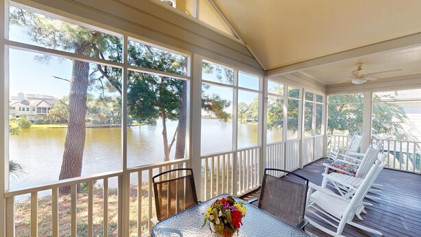 A covered patio with a table and chairs, large windows overlooking a body of water and trees, and a ceiling fan. - Beautiful 2BR 2BA Lakeside Villa with Lake Views (Pawleys Island)