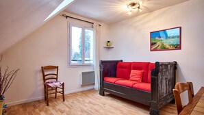 Ein Zimmer mit Holzboden, einem roten Sofa, einem Holzstuhl, einem Esstisch, einem Fenster mit Vorhängen, einer an der Wand befestigten Ablage mit einer Pflanze und einem gerahmten Bild.