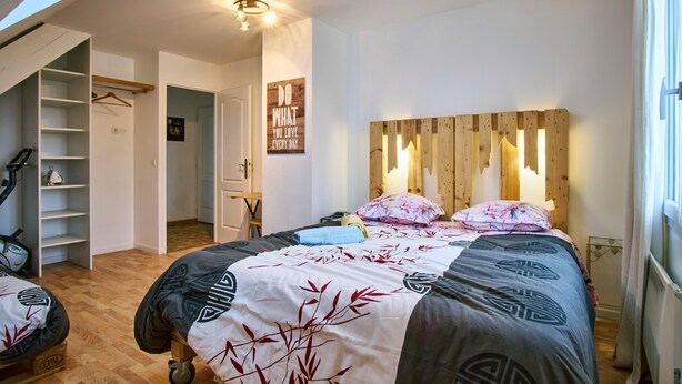 Ein Schlafzimmer mit einem Bett, einem hölzernen Kopfteil, einer Wandleuchte, einem Schrank mit Regalen und einer Tür zu einem anderen Raum.