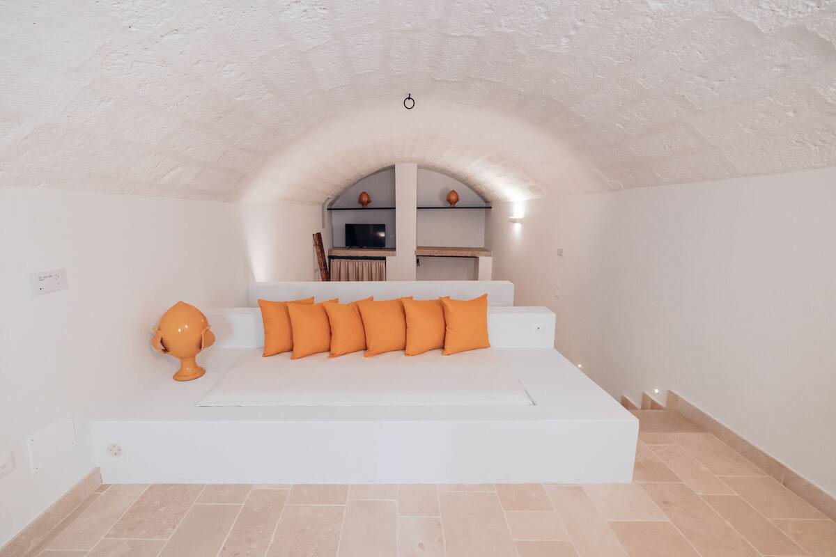 Un dormitorio moderno con un sofá blanco, cojines anaranjados, un televisor y un jarrón decorativo.