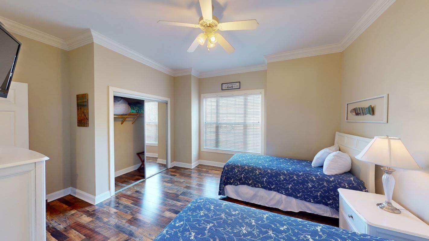 A bedroom with two beds, a ceiling fan, a bedside table with a lamp, a window with blinds, and a door leading to another room.