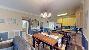 A spacious dining area with a wooden table and chairs, a chandelier, and a kitchen in the background. - Escape to Green Haven a Golfer's Delight (Pawleys Island)