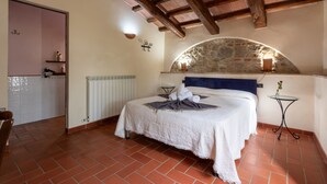 Un dormitorio con un arco de piedra, una cama con colcha blanca, una mesita con un jarrón y un radiador.