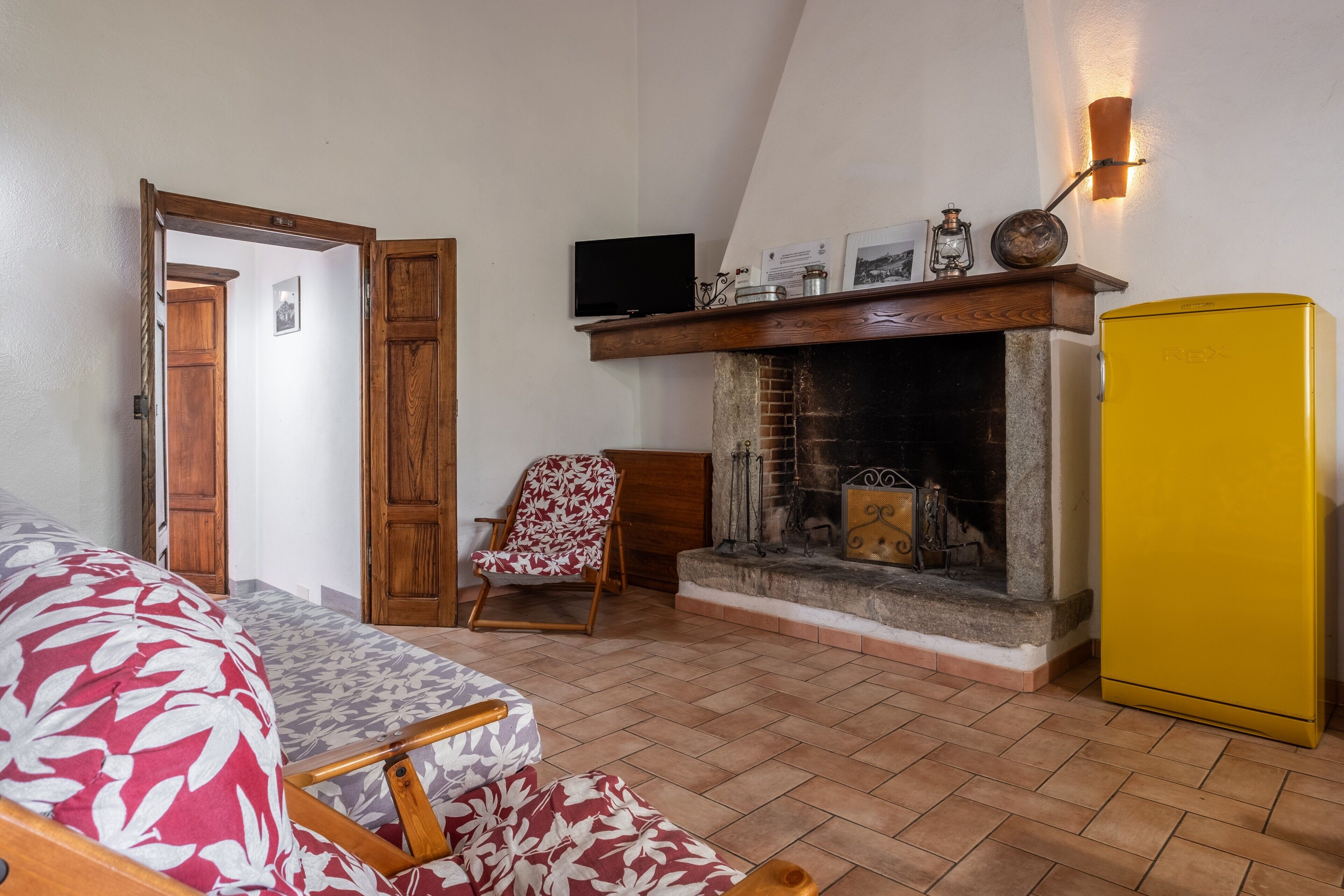 Ein Zimmer mit Kamin, einem gelben Kühlschrank, einer Holztür, einem gemusterten Sofa und einem Sessel.