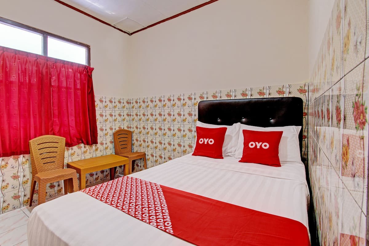 A hotel room with a bed, two chairs, a small table, floral wallpaper, and a window with red curtains.