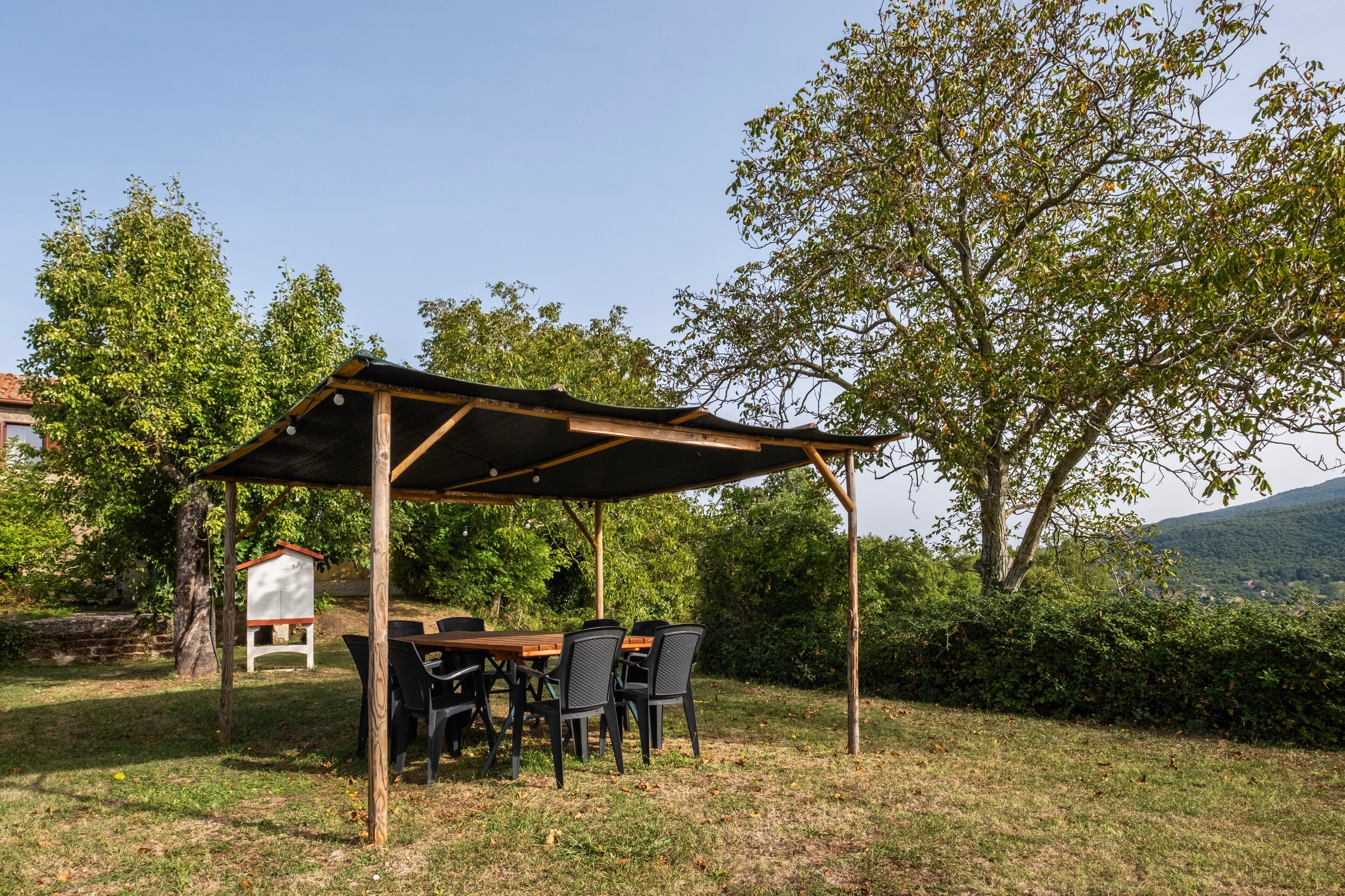A covered outdoor dining area with a table and chairs, surrounded by trees and a view of a hill.