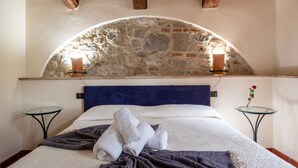 Quarto com arco de pedra, cabeceira azul, cama com roupa de cama branca, duas mesas de cabeceira e uma pequena mesa com um vaso de flores.
