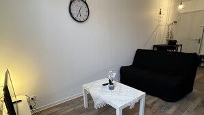 A living room with a white coffee table, a black sofa, a clock on the wall, and a television on a stand.