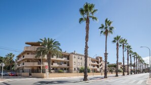 Un immeuble à plusieurs étages doté de balcons, des palmiers bordant la rue et un ciel d’un bleu limpide.