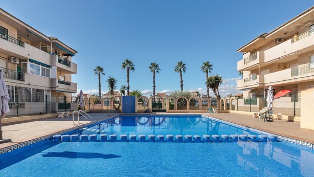 Une piscine dont le bord est revêtu de carreaux, entourée de bâtiments dotés de balcons et de palmiers.