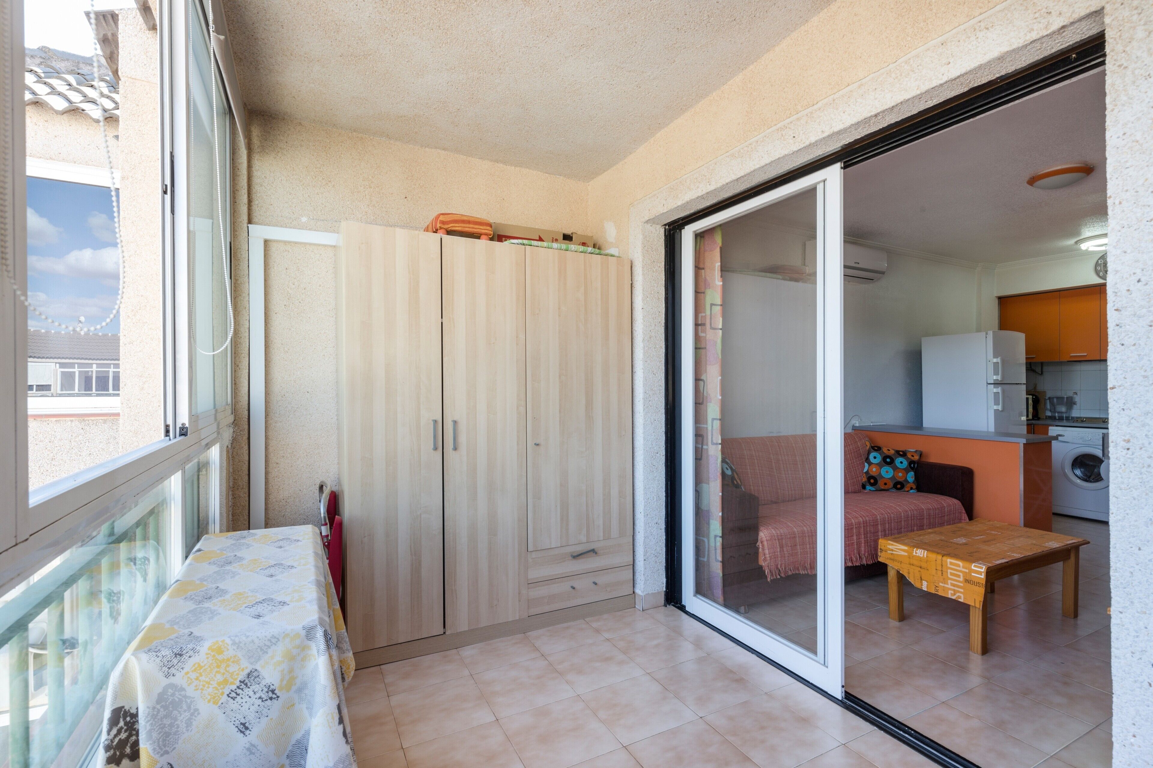 Una habitación con cama, una mesa, un armario y un ventanal corredero que da a una cocina.