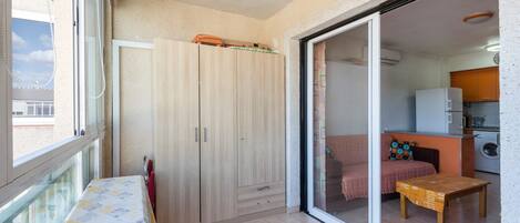 Una habitación con cama, una mesa, un armario y un ventanal corredero que da a una cocina.