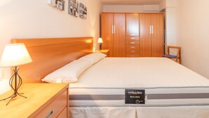 Una cama bien hecha con un cojín, una lámpara de noche, una mesita de madera y un armario de madera con varios cajones, junto a una silla.