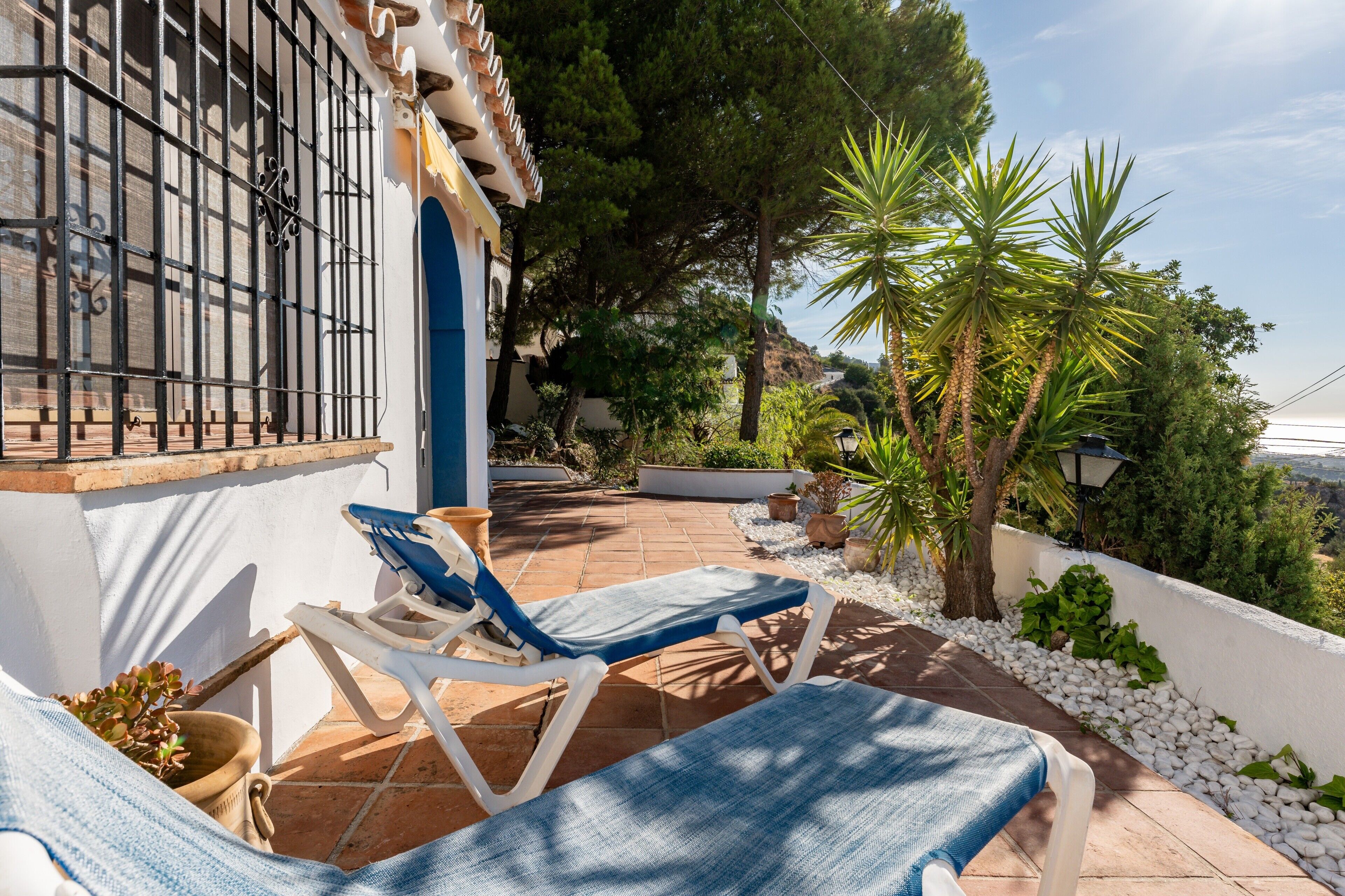 Una terraza con dos tumbonas, una pared blanca con un ventanal y un jardín con árboles y plantas.