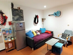 Un espace de vie compact comprenant un canapé, une petite table, un réfrigérateur, une guitare et un luminaire fixé au mur.