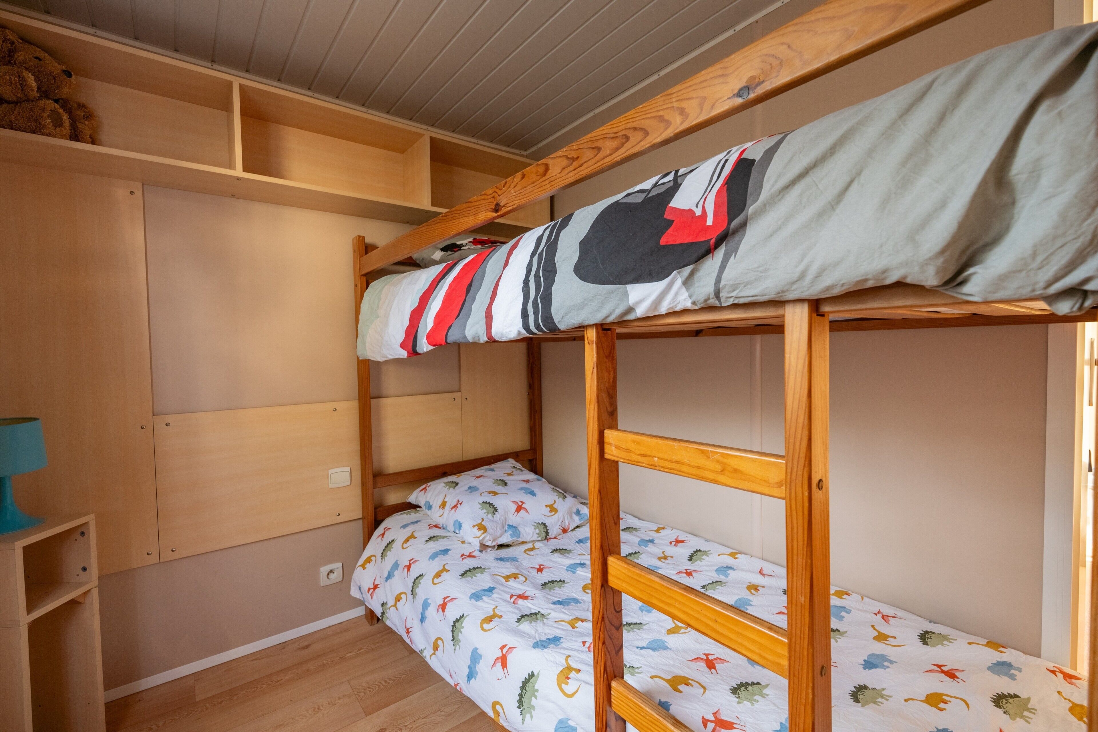 A bunk bed with a patterned bedspread, a wooden shelf unit, and a small table.
