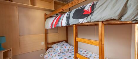 A bunk bed with a patterned bedspread, a wooden shelf unit, and a small table.