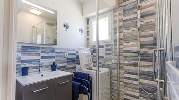 Une salle de bain moderne avec une douche à l’italienne en verre, un lavabo blanc doté d’un distributeur de savon bleu et un mur recouvert de carreaux.