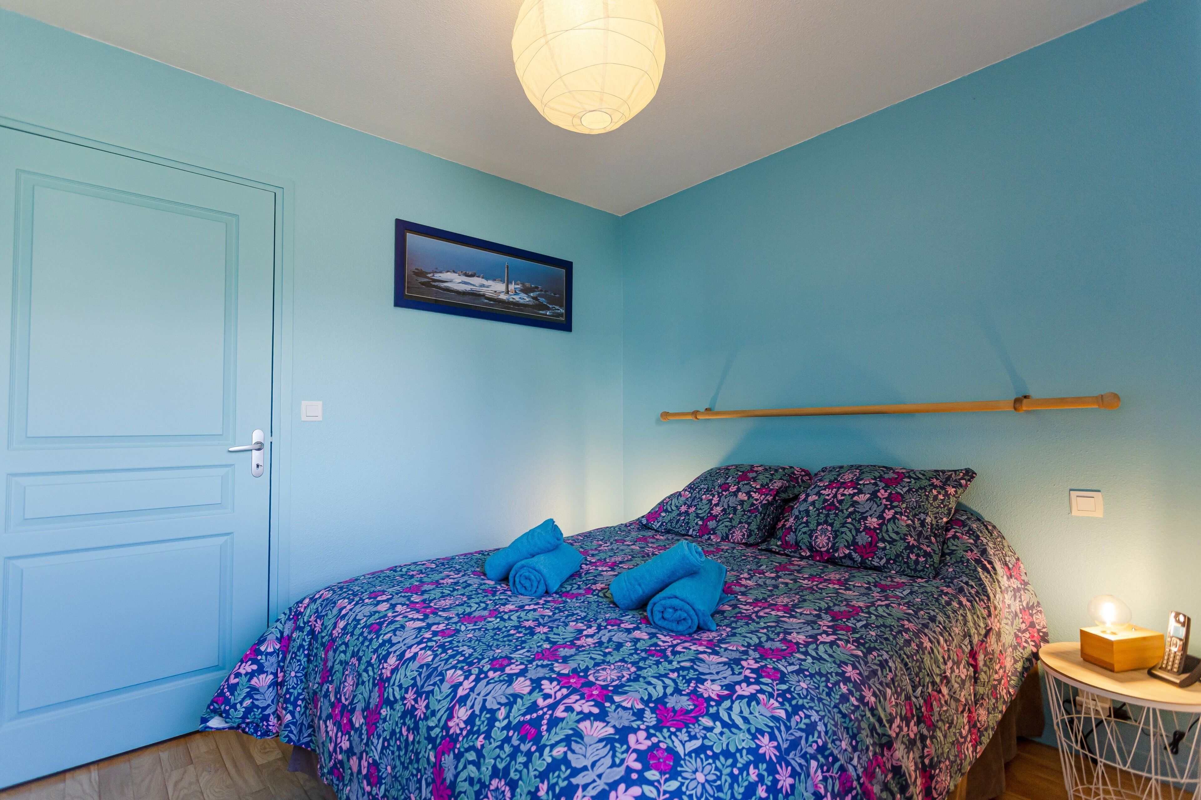 A bedroom with a bed, pillows, a framed picture, a bedside table with a lamp, and a door.
