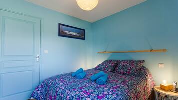 A bedroom with a bed, pillows, a framed picture, a bedside table with a lamp, and a door.