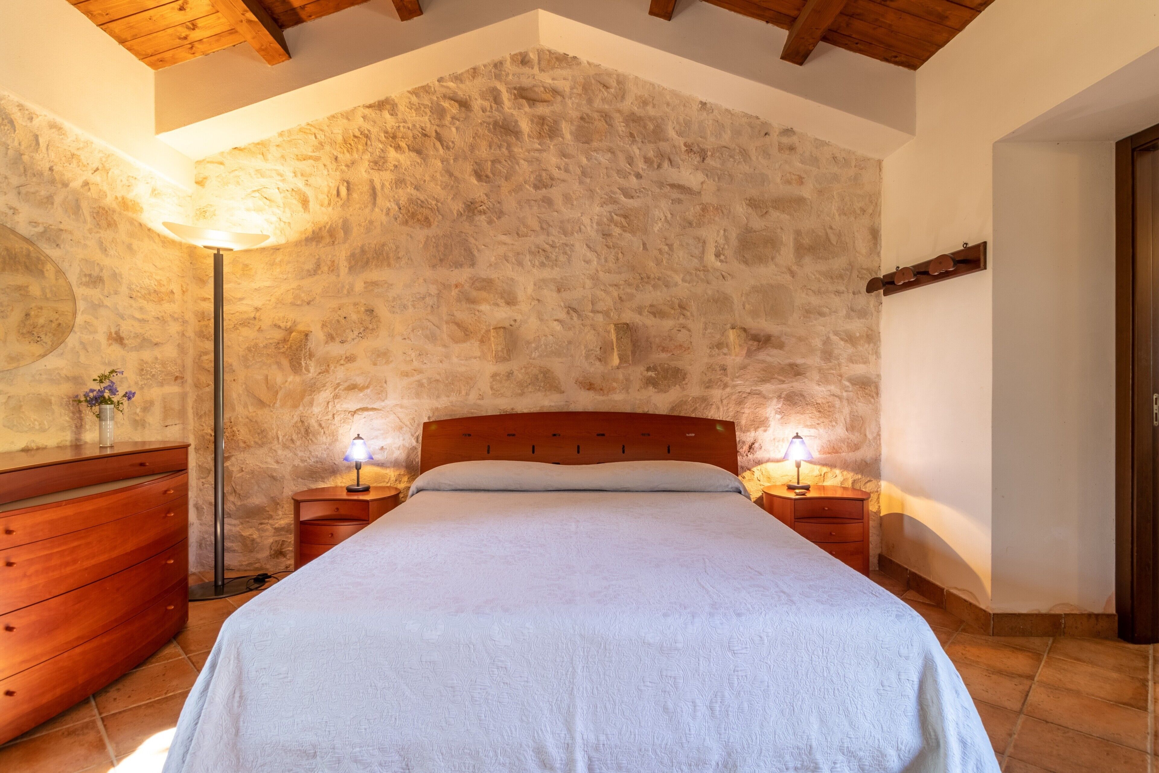 Een slaapkamer met een stenen muur, een houten bedframe, twee nachtkastjes met lampen en een houten dressoir.