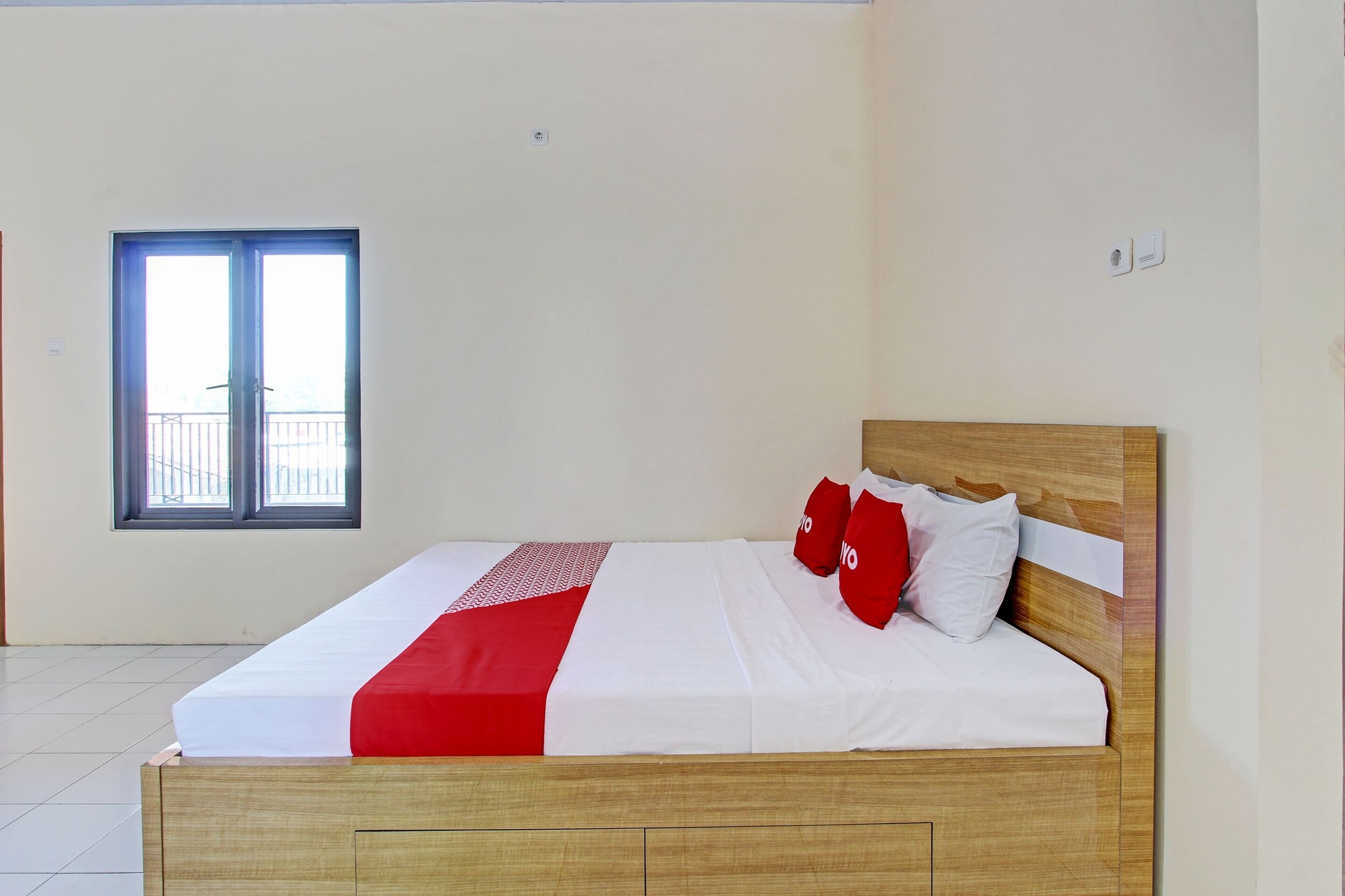 Een net opgemaakt bed met een houten hoofdbord, witte beddengoed en rode accenten. Een raam met uitzicht op een balkon is zichtbaar.