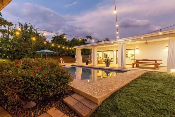 A backyard with a swimming pool, a covered patio, and string lights.