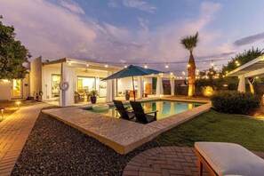 Zona de piscina con sillones de descanso, un espacio de estar cubierto y luces colgantes.