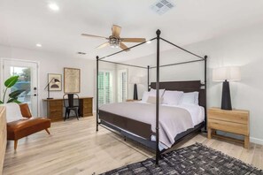 A bedroom with a four-poster bed, a desk, a chair, a bedside table, and a ceiling fan.