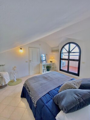 Een slaapkamer met een groot bed, een raam, een deur en een klein tafeltje met een lamp.