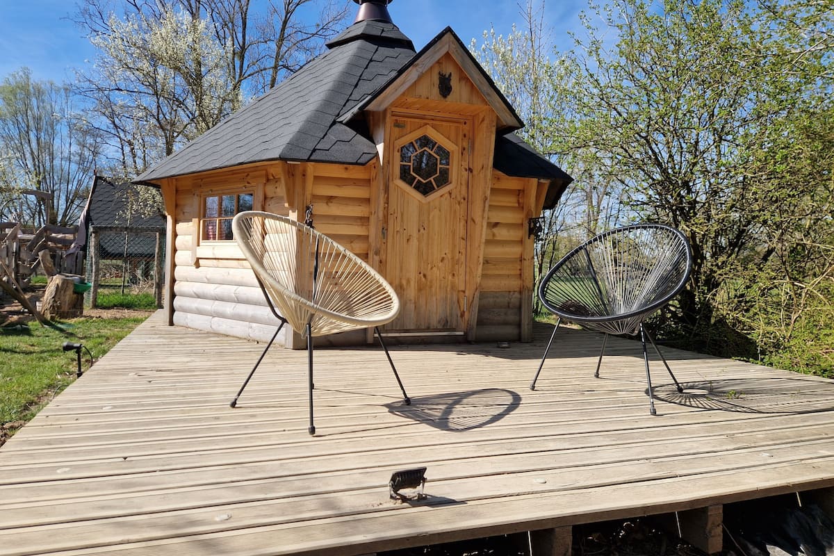 Een houten blokhut met een schuin dak, een schoorsteen en een houten deur met een raam. Twee rieten stoelen staan op een houten terras voor de blokhut.