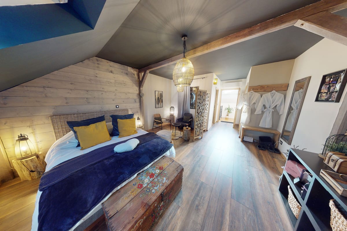 Een gezellige slaapkamer met een groot bed, een houten bankje, een kleine tafel en een stoel. Er is een gang die leidt naar een badkamer met een spiegel en een handdoekhouder.