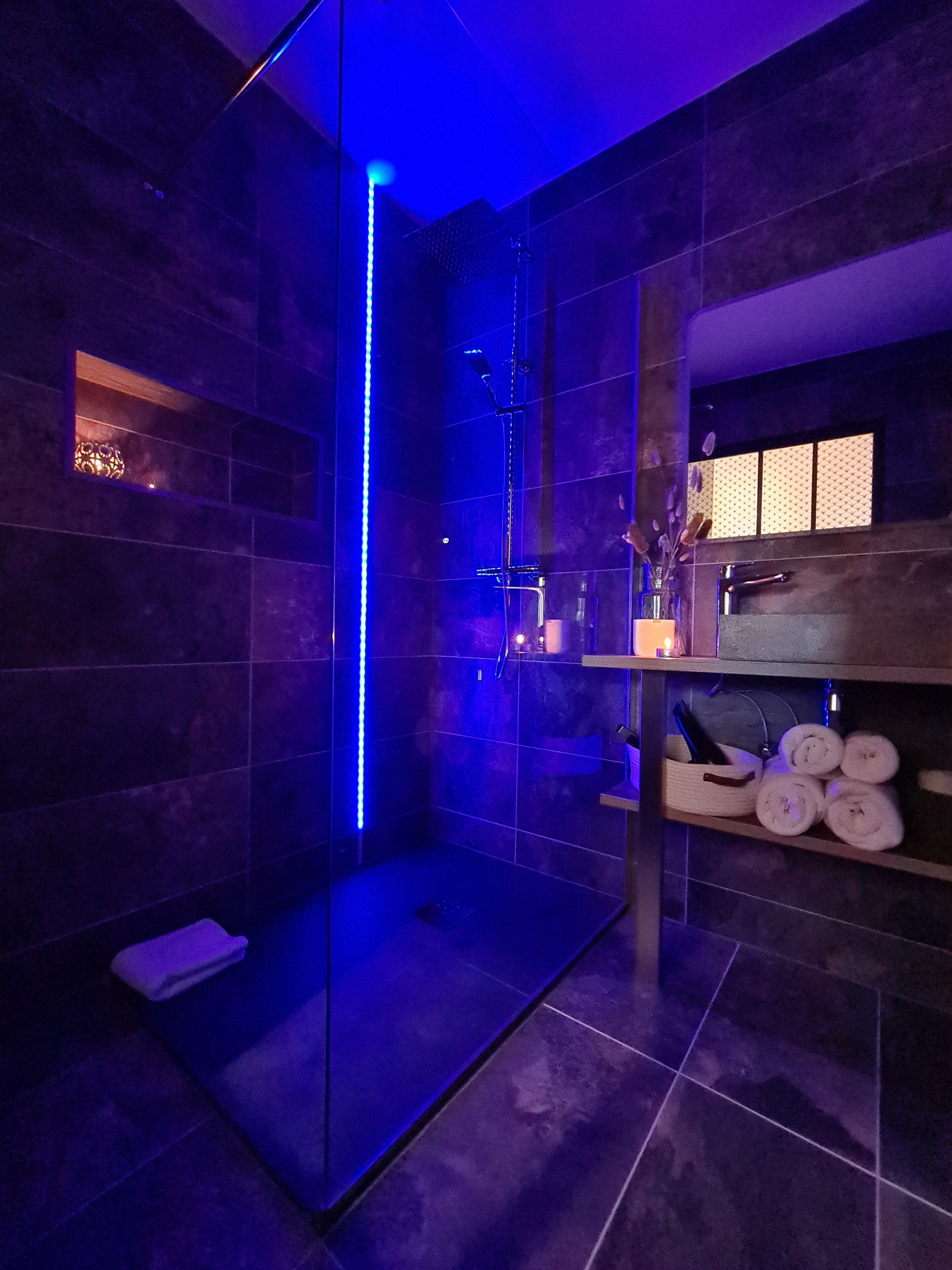A modern bathroom with a glass-enclosed shower, dark tiled walls, and a shelf with towels and toiletries.
