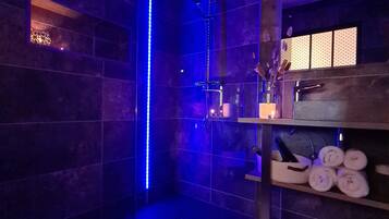 A modern bathroom with a glass-enclosed shower, dark tiled walls, and a shelf with towels and toiletries.