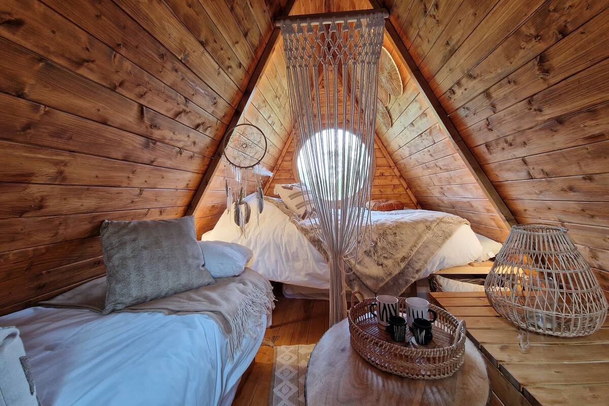 Een knus slaapkamer onder dak met houten wanden, een hangende dromenvanger, een bed met kussens, een geweven mand met mokken, en een geweven stoel.