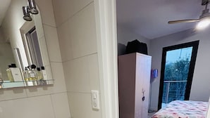 Une chambre avec un plancher en bois, un lit recouvert d’une couvre-lit à motifs floraux, une armoire blanche et une salle de bain avec un lavabo et un miroir.
