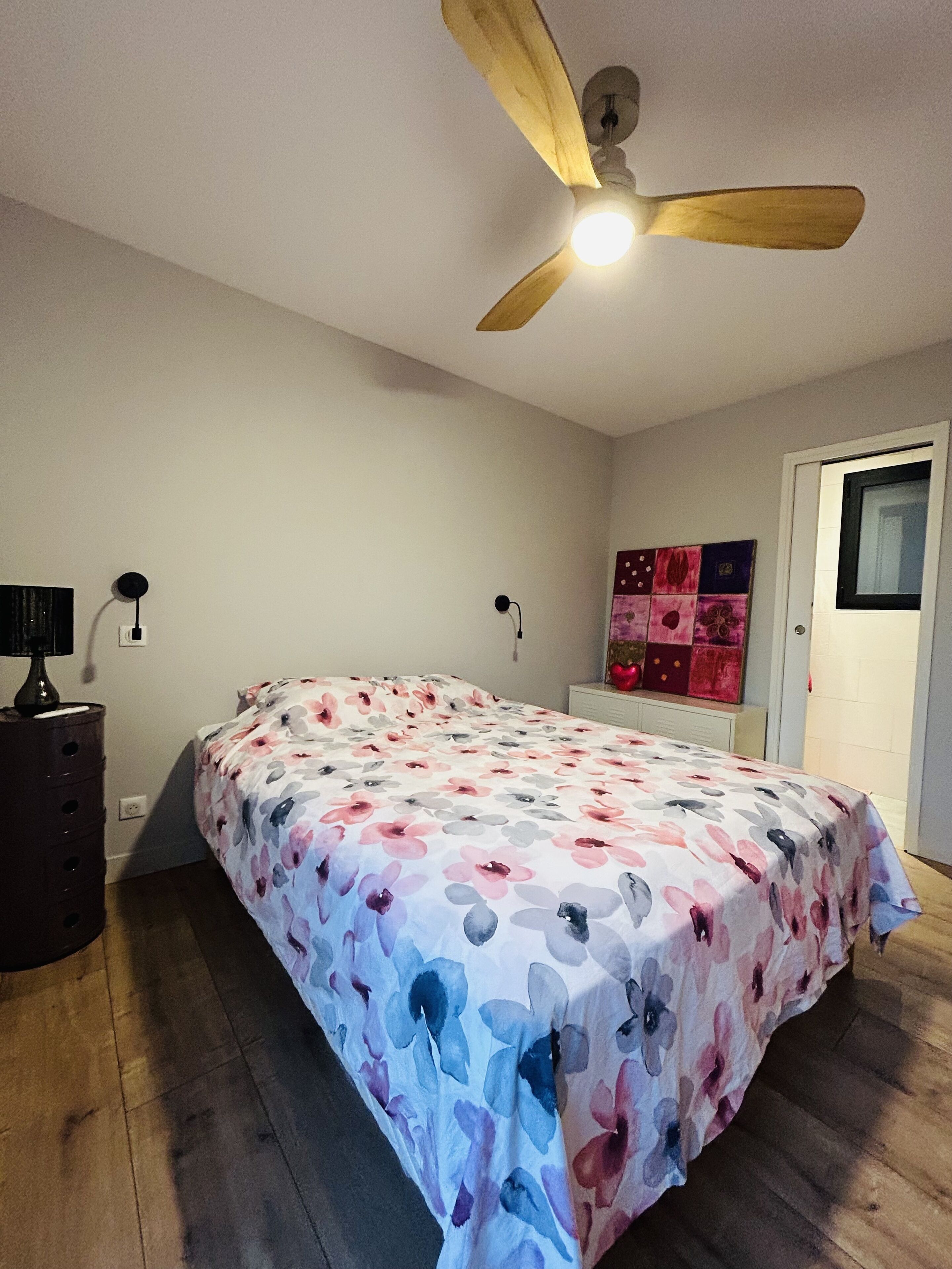 Ein Schlafzimmer mit einem Bett, einem Deckenventilator, einer Kommode, einem Nachttisch und einem Wandbild.
