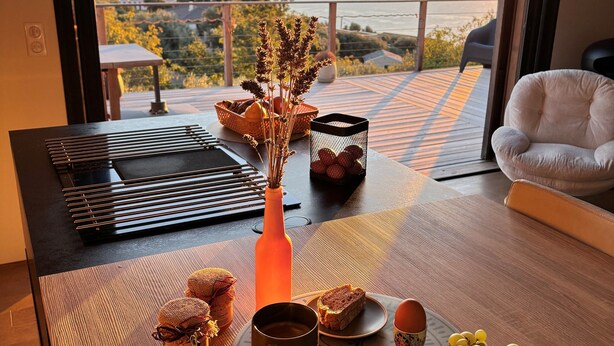 Une table à manger avec vue sur le coucher de soleil, un vase contenant des plantes séchées, une bouteille, du pain et une assiette de raisins.