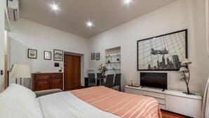 Ein Hotelzimmer mit einem Bett, einem Schreibtisch mit Stuhl, einem Fernseher und einem Stadtbild im Bilderrahmen an der Wand.