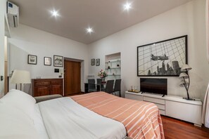 A hotel room with a bed, a desk with a chair, a television, and a framed cityscape picture on the wall.