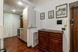 A compact kitchen with white cabinets, a stove, and a refrigerator. There are framed pictures on the wall and a wooden cabinet with drawers.