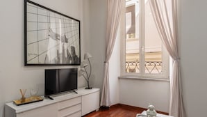 Une pièce comprenant un grand tableau encadré, un téléviseur à écran plat, une petite table sur laquelle est posée une lanterne blanche, et une fenêtre avec des rideaux.