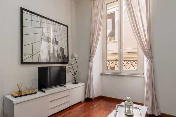 A room with a large framed picture, a flat-screen TV, a small table with a white lantern, and a window with curtains.