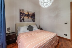 Un lit bien fait, recouvert d’une couvre-lit à rayures, une table de chevet avec une lampe, un tableau représentant un paysage urbain accroché au mur, et un lustre moderne.