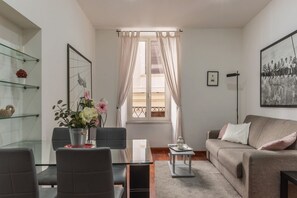 A modern living room with a glass dining table, grey sofa, and a large window with sheer curtains.